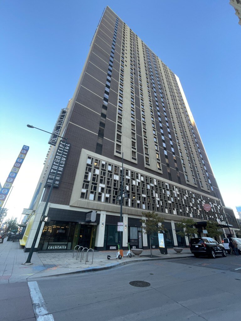 Looking up at the tall conference hotel tower in downtown Denver from the street corner, with shopfronts and a vertical ALL TOGETHER sign at its base.