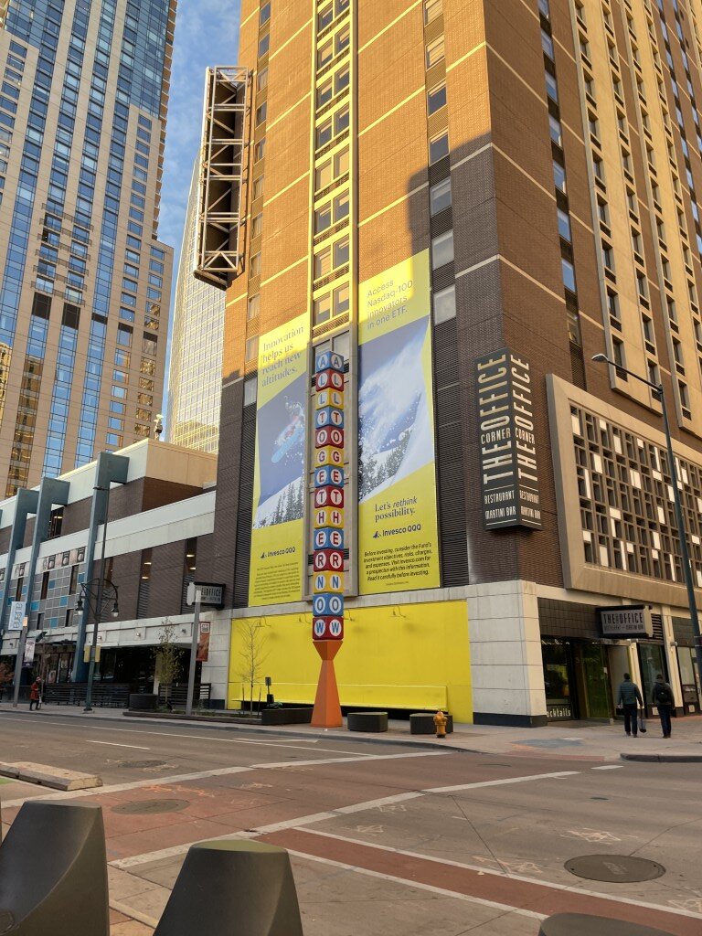 The conference hotel seen from across the street, with a column of coloured letter tiles spelling ALL TOGETHER and a bright yellow wall at ground level.