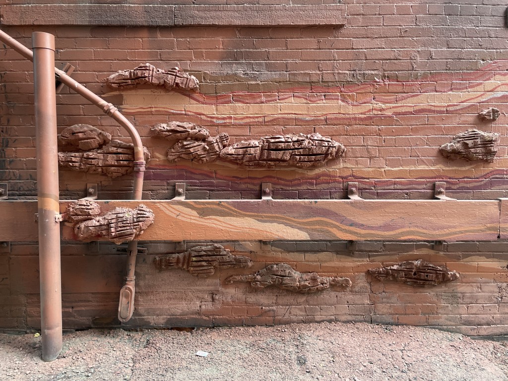 A downtown Denver alley wall painted with flowing sandstone-coloured stripes, with sculpted rock formations projecting out of the brickwork.