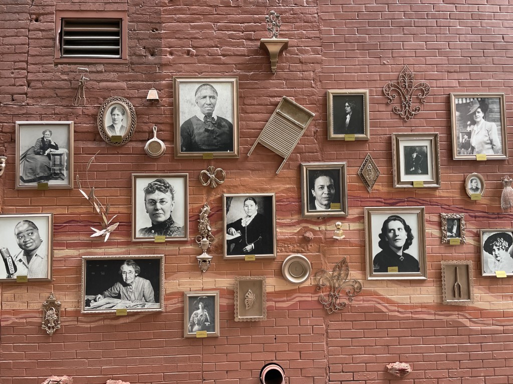 A pink brick wall hung with framed black-and-white portraits of women alongside vintage household objects, arranged as a gallery-style mural.