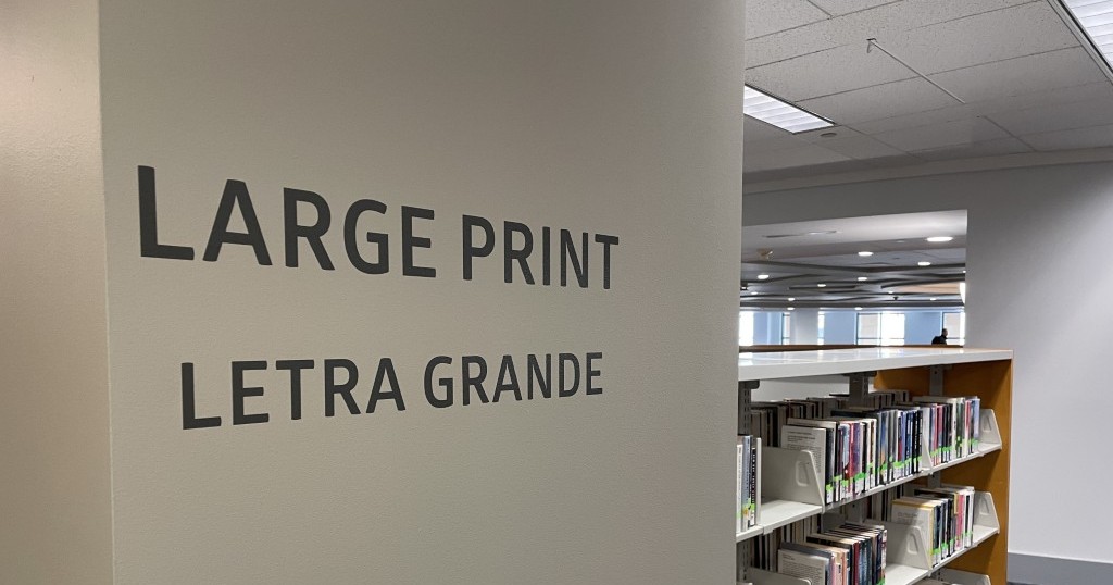 A library pillar with large dark lettering reading LARGE PRINT and LETRA GRANDE, with shelves of large-print books visible behind it.