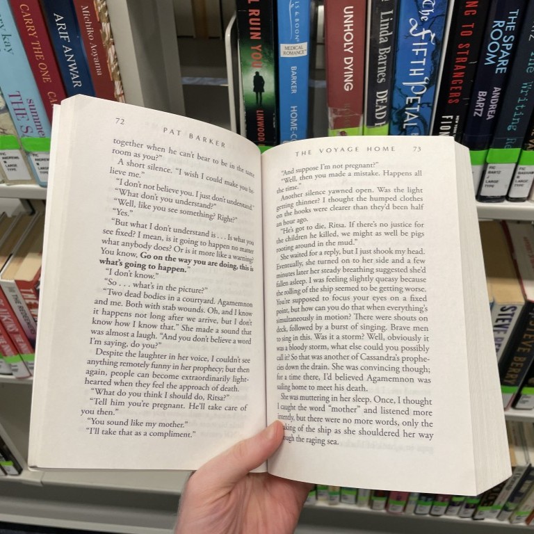 A large-type paperback held open in one hand in front of shelves of hardcover novels.