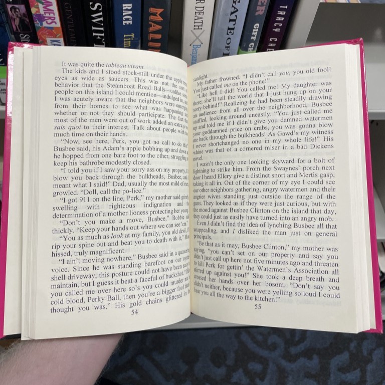 A large-type hardcover held open, its text set widely across both pages, in front of a shelf of paperbacks.