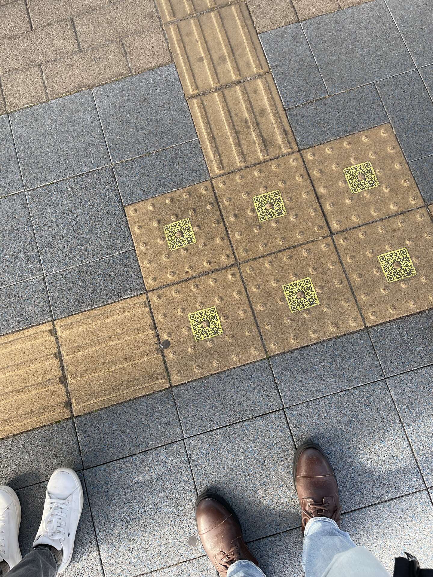 Yellow tactile paving tiles outdoors, each dotted warning tile carrying a printed QR code at its centre, with three pairs of feet standing around them.