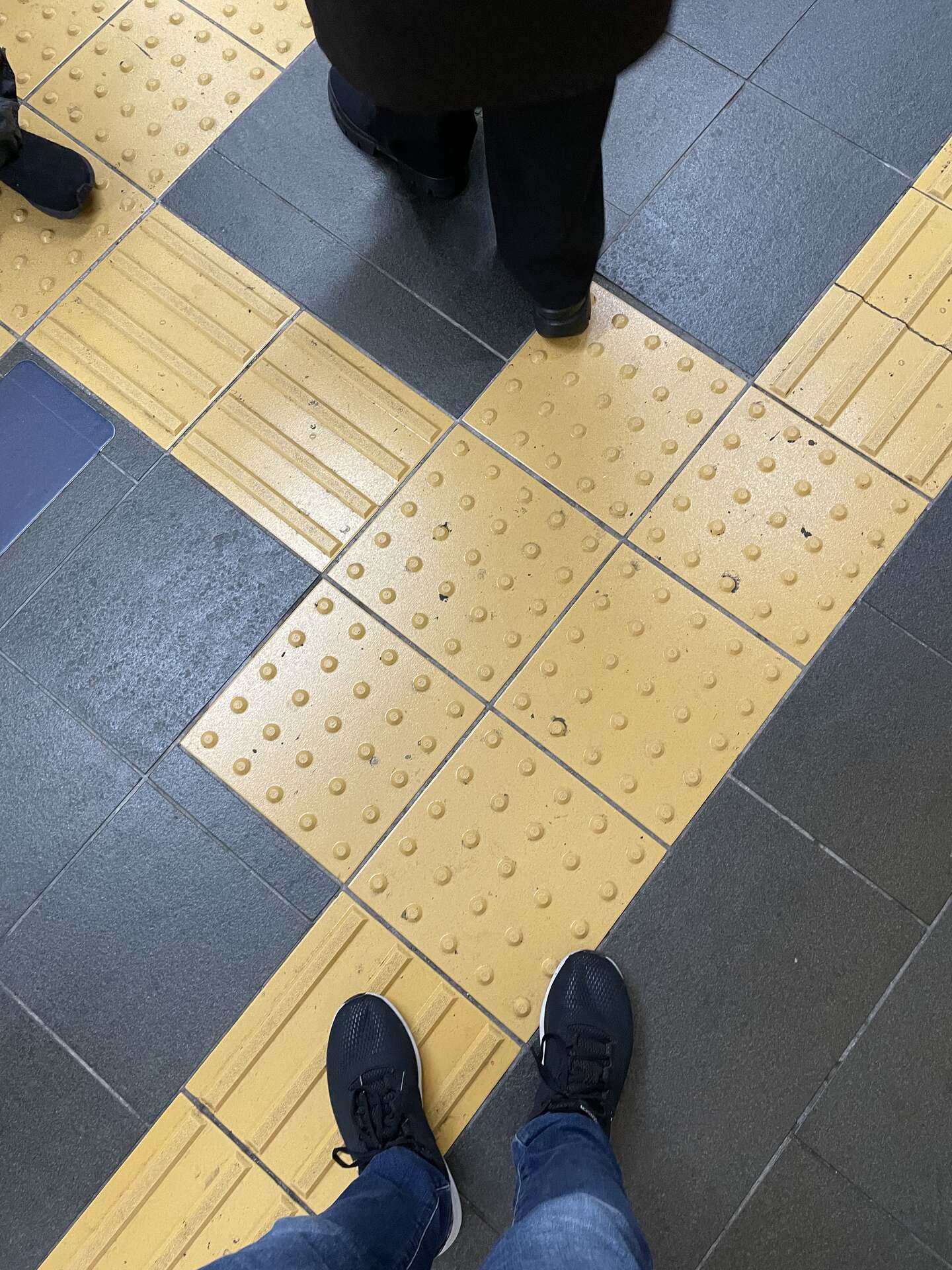Looking down at feet standing on yellow tactile paving, where dotted warning tiles meet ribbed directional tiles against grey stone.