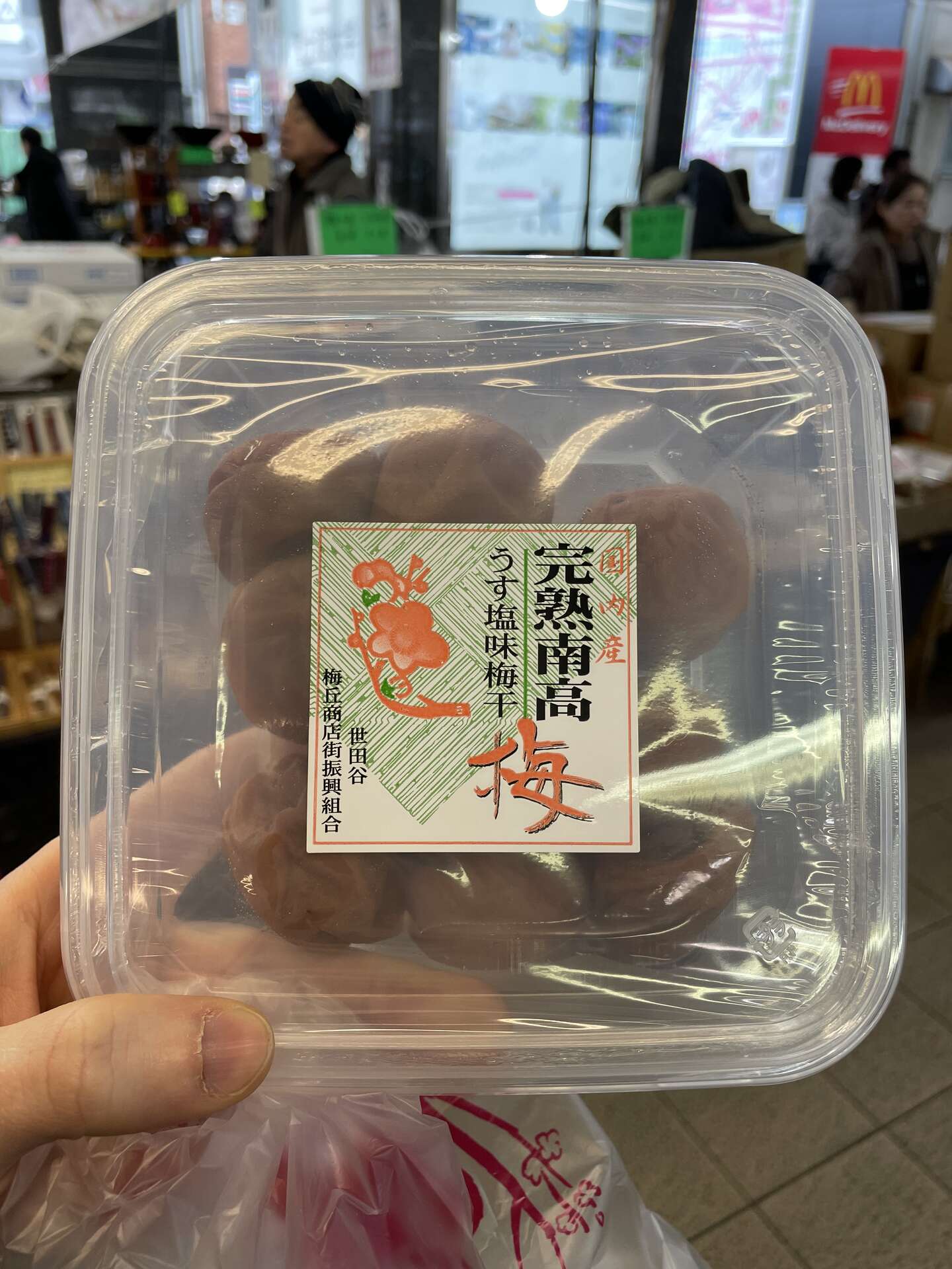 A clear plastic tub of umeboshi held up to the camera, its label reading 完熟南高梅 with a red plum blossom illustration.