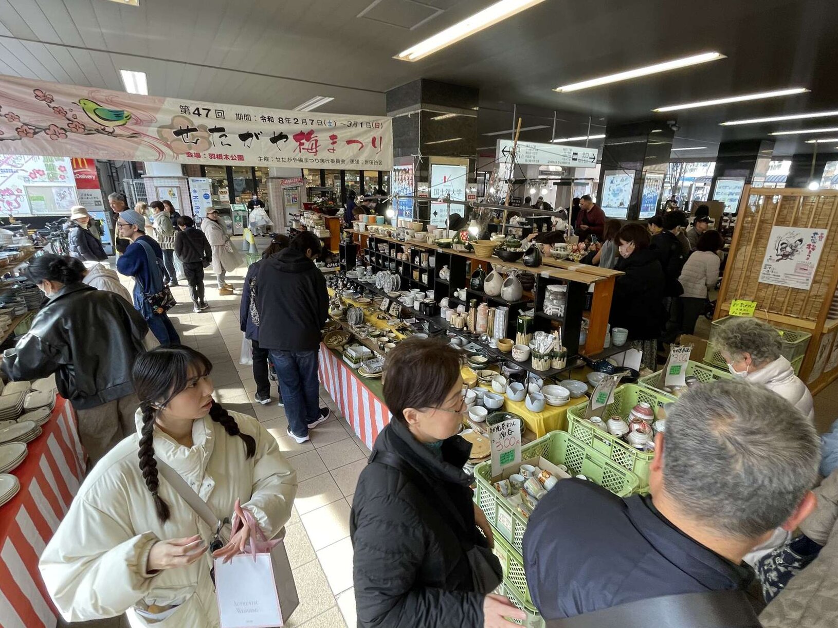 A busy market inside the station concourse, with stalls of ceramic bowls and tableware under a banner for the 47th Setagaya Plum Festival.