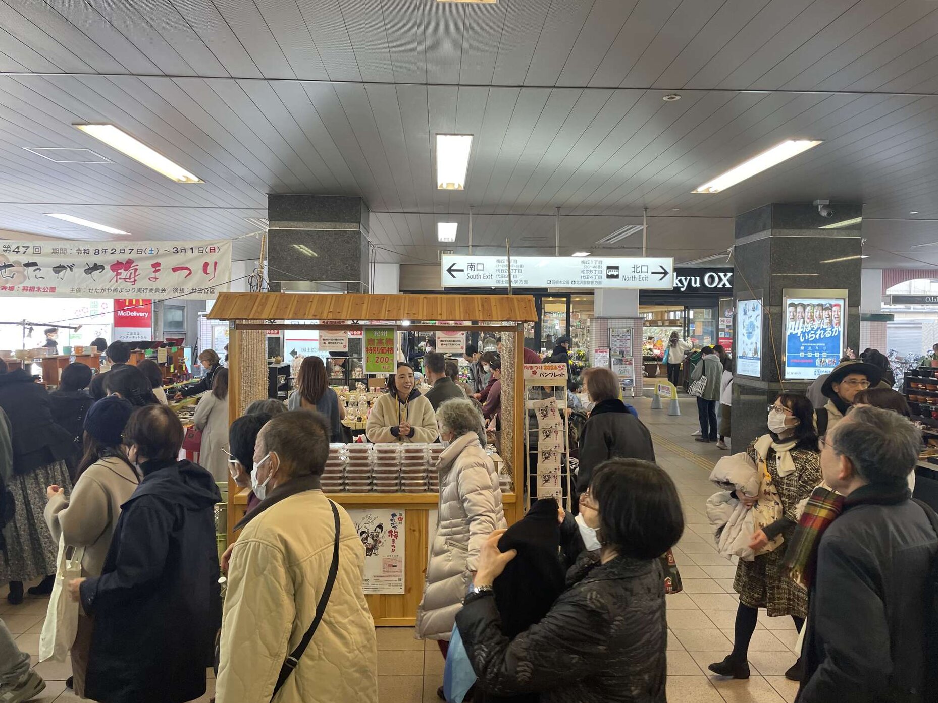 Shoppers crowding around a wooden stall selling boxed umeboshi in the station concourse, beneath signs for the south and north exits.