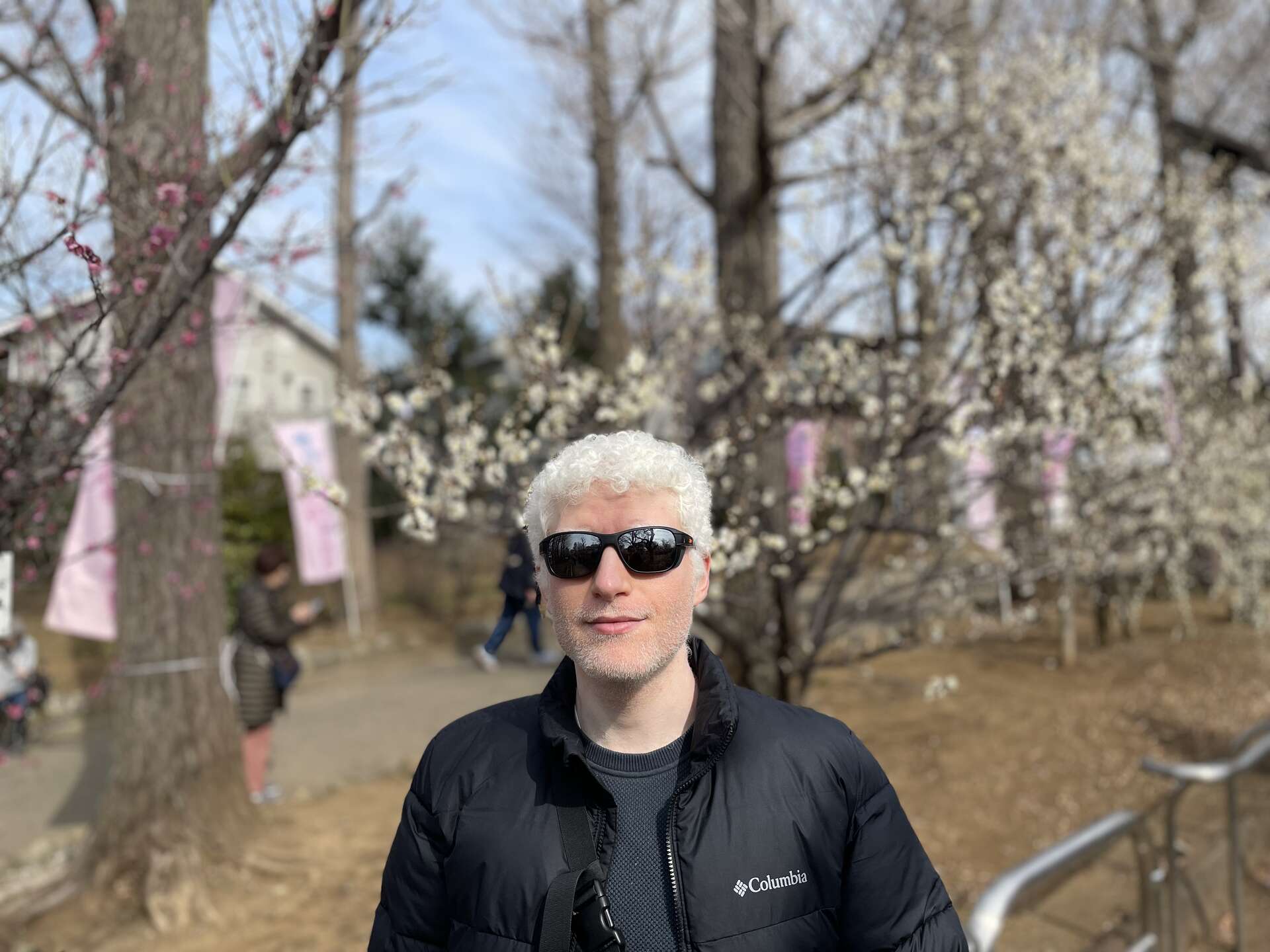 Yotam in sunglasses and a black jacket smiling at the camera on a park path, with white and pink plum trees and festival banners behind him.