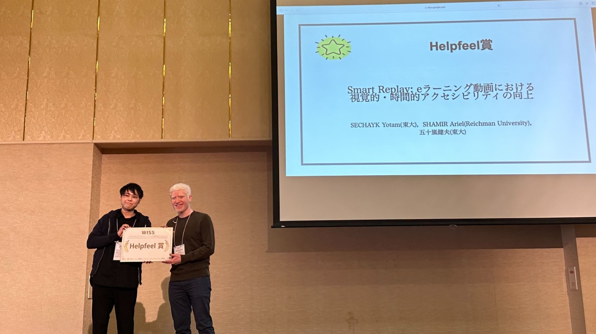 Yotam and a Helpfeel representative stand on the WISS 2023 stage holding the award certificate between them, with the Helpfeel賞 slide for the Smart Replay paper projected on the screen behind.